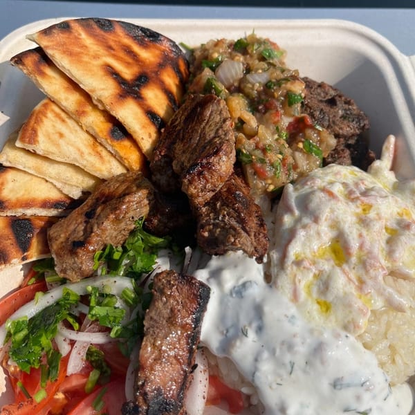 A Mediterranean dish with grilled meat, pita bread, rice, salad, and various dips in a takeout container.