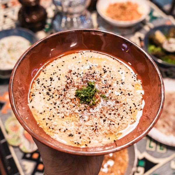 A hand holding a bowl of Mediterranean hummus, garnished with sesame seeds, paprika, and parsley.